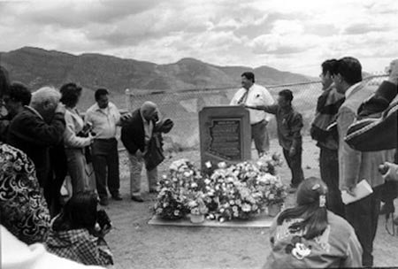 A group of people gathered around a memorial plaque with flowers in a mountainous area.